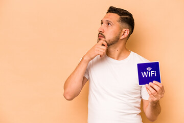 Hispanic man holding wifi placard isolated on beige background looking sideways with doubtful and...