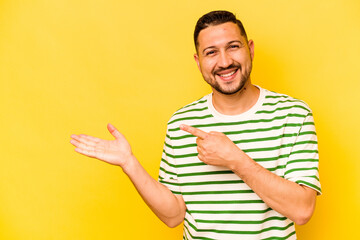 Young hispanic man isolated on yellow background excited holding a copy space on palm.