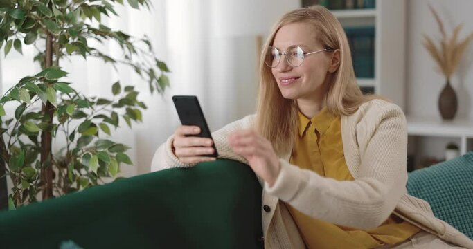Attractive mature woman in eyeglasses sitting on comfy couch and surfing web pages on modern smartphone. Beautiful blonde relaxing at home with gadget in hands.