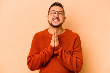 Young hispanic man isolated on beige background holding hands in pray near mouth, feels confident.