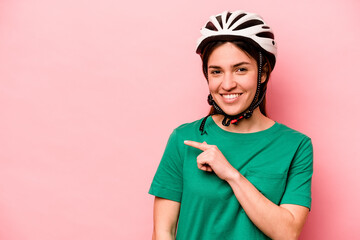 Young caucasian woman wearing helmet isolated on pink background smiling and pointing aside, showing something at blank space.