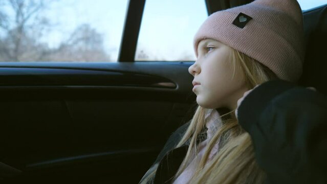 Beautiful female kid with long blonde hair looks outside through the window in the backseat of a moving car. Small sad girl rides on modern SUV and watches at nature through the glass of auto. Slow mo