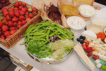 Assortment of greens, berries, cheese and honey on the table. Micro green leaves, juicy pea pods, strawberries in a wicker basket, assorted cheese with raspberries and blackberries, honey, prosciutto.