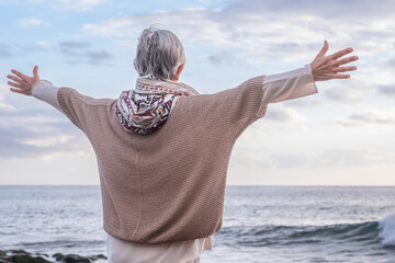 Rear view of caucasian senior woman standing at the beach with outstretched arms looking at horizon...