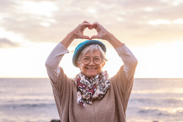 Portrait of caucasian senior woman standing at sunset light at the beach gesturing heart shape with hands looking at camera. Happy smiling elderly lady enjoying sea vacation