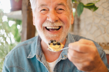 Close up on happy bearded senior man eating a fruit cake in coffee shop enjoying break or breakfast and looking at camera