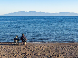 Web banner rear view of father and child sitting in deckchairs at beach on sunny winter day. Single parenting. Happy family togetherness. Mid aged adult male and young kid. Background with copy space.