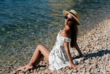 A beautiful young woman in a hat, glasses and a light dress is sitting with her back to the ocean against the background of huge rocks on a sunny day. Tourism and tourist trips.