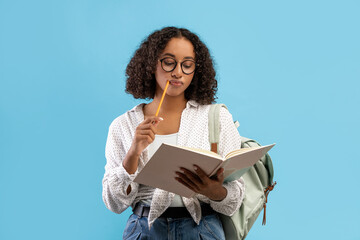 Thoughtful African American female student reading book, thinking over home assignment on blue studio background