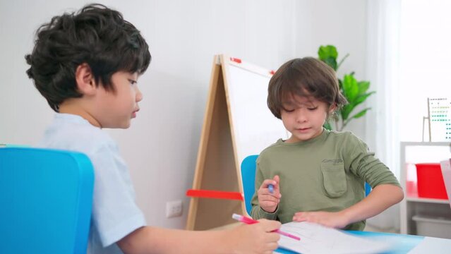 Two handsome boys sitting in the kindergarten school classroom and drawing or painting on the paper with color pencil. Kid help friend to finish drawing. Learning, enjoy, education, playing concept