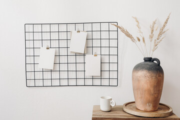 Black metal mesh noticeboard, bulletin board with blank memo cards mockups. Elegant home office interior concept. Vase with dry lagurus grass and cup of coffe on wooden table. White wall background.