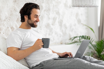 Handsome young guy using laptop in bed, drinking coffee