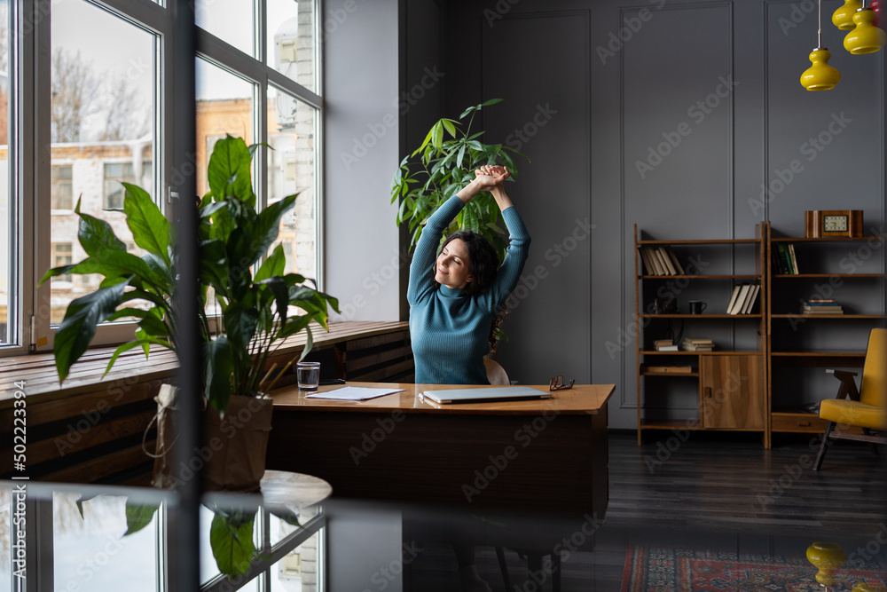 Wall mural young relaxed businesswoman sitting at office desk, relaxing with hands over head, looking out windo