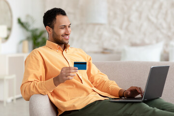 Smiling man using pc and credit card sitting on couch