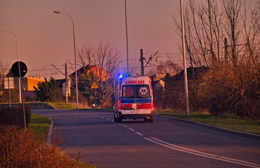 Ambulans na sygnale ( włączone migające lampy " koguty " ) jadący miejską ulicą , przy zachodzącym słońcu .