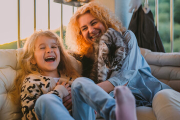 a little, happy girl, a child with a smile plays on a summer, spring vernada at home with mom and a cat at sunset. family weekend evening. cat, pets as a family member.