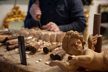 Master woodcarver at work. Wood shavings, gouges and chisels on the workbench. Angel wood carved in the foreground.. - obrazy, fototapety, plakaty