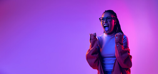 Portrait of excited young girl wearing in cotton shirt shouting isolated on purple background in neon light. Concept of beauty, art, fashion, youth and emotions