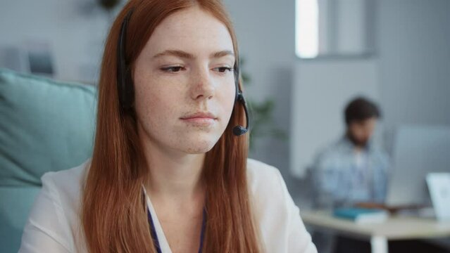 Close-up portrait of young redhead woman in modern office. Female Caucasian secretary in office clothes with headphones and microphone solving client's problem in customer support center. Blured