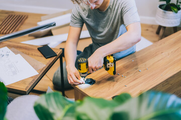 A young man with an electrical screwdriver assembles a tv stand console according to instructions in his new house. Man assembling furniture at home using a cordless screwdriver. Selective focus.