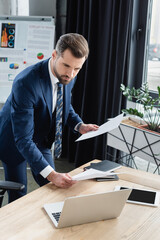 Naklejka premium businessman in suit holding papers and looking at laptop in office.