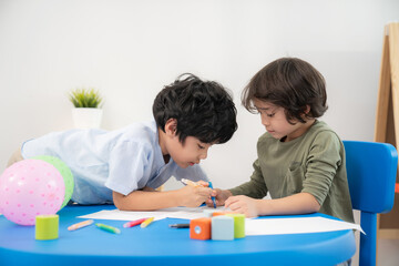 Two handsome boys sitting in the kindergarten school classroom and drawing or painting on the paper with color pencil. Kid help friend to finish drawing. Learning, enjoy, education, playing concept