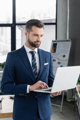 economist in suit using laptop while standing in blurred office.