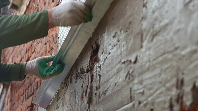 A bricklayer levels the plaster on a brick wall with a spatula. Plastering cement at wall for renovation house. Selective focus. Putty brick wall background. Plasterer smoothing plaster on wall.