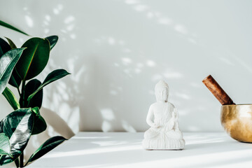 Decorative white Buddha statuette, green plant and tibetian singing bowl on the white background with sun light shadows. Meditation and relaxation ritual. Exotic massage. Minimalism. Copy space. - obrazy, fototapety, plakaty