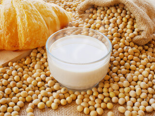 A hot cup of soy milk on lots of soy beans and sack of soy beans and croissant on wooden cutting board