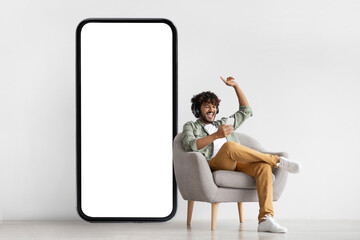 Joyful indian guy sitting in armchair with smartphone and headset - Powered by Adobe