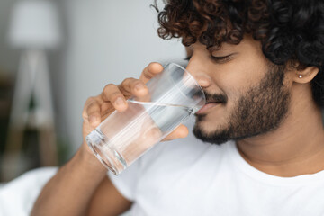 Happy curly young man taking glass of water in bed