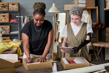 Happy girl with partial arm using tablet while her colleague packing parcel with folded clothes and...