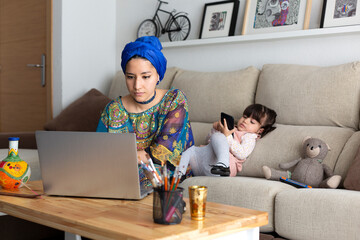 Young Muslim woman working at home with her laptop while her young daughter plays with a mobile phone. Single parent family.