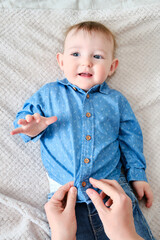 A mother woman wears a blue shirt to a baby boy. Mom dresses a happy child in clothes on the bed.