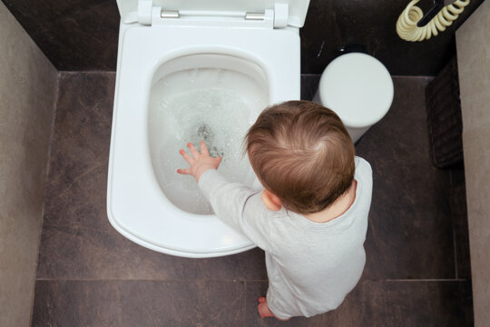 Toddler baby boy is playing in the toilet room with water flush in the white toilet bowl. Child plays with white toilet seat cover