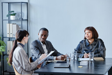 Minimal shot of business team at meeting table in office against grey wall, focus on smiling African American man talking to female colleagues