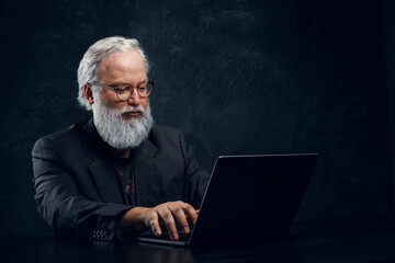 Studio shot of old man dressed in black suit using laptop against dark background.