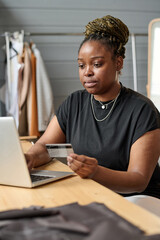 Young modern female consumer paying by credit card for ordered online goods while sitting in front of laptop and looking at screen