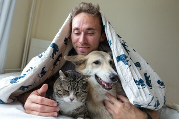 Pleased young man lying in bed with dogs and cat