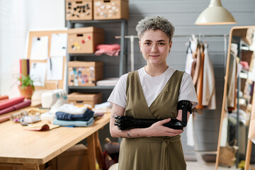 Young successful female tailor with partial arm standing against her workplace with table,...