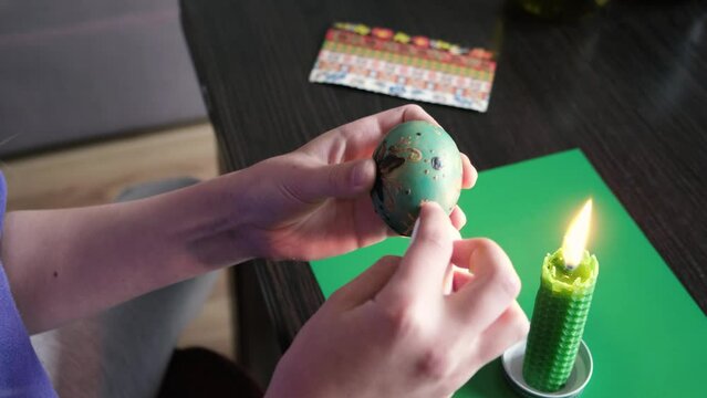 With the help of a candle, the girl melts the wax and removes it with a napkin from an Easter egg. Close-up.