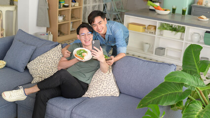 Young smiling gay couple taking a selfie with healthy food in the living room at home, LGBTQ and...