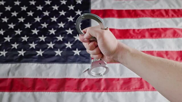 caucasian hand holding and turning silver metal handcuffs over blurry United States flag