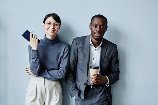 Candid Waist Up Portrait Of Two Business People Caucasian And African American Smiling At Camera While Standing Against Simple Grey Background In Office