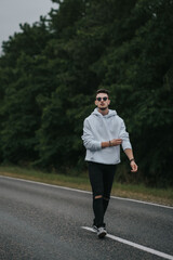 A young handsome guy with a small beard in a white jacket and dark glasses is walking along the road