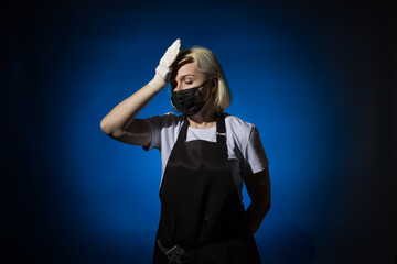 A gardener woman in a black apron, gloves in a protective mask raised her hand to her head on a dark background.