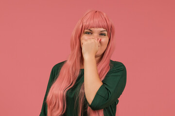 a woman with long pink hair in a green dress pinched her nose from the smell on a pink background