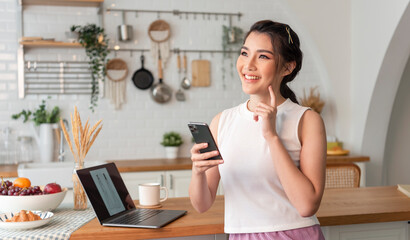 Young woman Asian happy and interested in thinking using mobile phone in kitchen room.