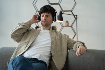 A young adult man is sitting on a comfortable sofa wearing wireless headphones enjoying listening to modern music and dancing. 